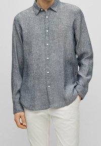 Knappkrage skjorta i ljusgrått tyg med en subtil textur, långa ärmar, klassisk krage och vita knappar. Vita byxor synliga.