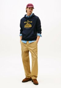 Marineblauer Kapuzenpullover mit gelber Grafik, darunter ein Jeanshemd, beigefarbene Cargo-Hosen und braune Loafers, vor einem schlichten weißen Hintergrund.