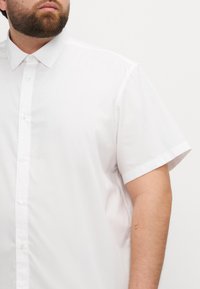 Cămașă albă cu mâneci scurte, cu guler cu nasturi, textură fină și croială normală; are nasturi în față și un design curat, minimalist.