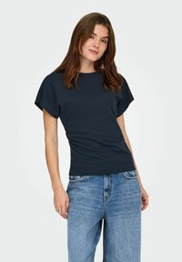 Jeune femme aux longs cheveux bruns portant un haut noir à manches courtes et un jean bleu ample, debout devant un fond uni.