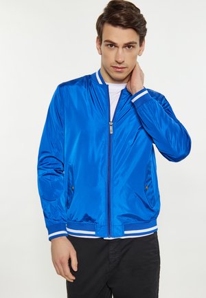 Jeune homme portant une veste zippée bleue brillante avec des bordures blanches, touchant son cou d'une main, debout devant un fond blanc uni.