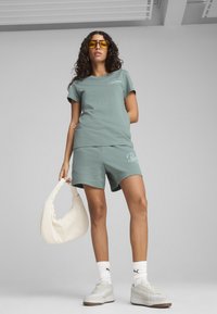 Camiseta de algodón verde claro con mangas cortas y logo, combinada con pantalones cortos a juego. Una bolsa de hombro blanca y unas zapatillas completan el look.