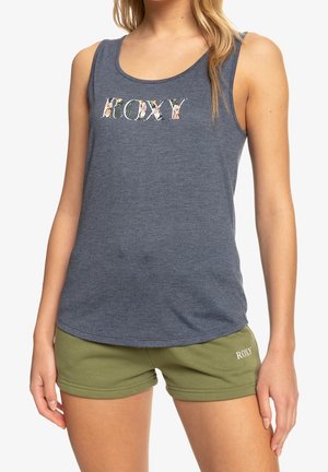 Vrouw draagt een mouwloos donkergrijs hemd met bloemen "ROXY" tekst en olijfgroene shorts met "ROXY" logo op het linkerbeen.