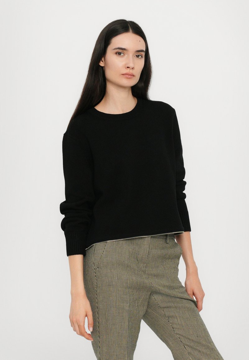 Black, round-neck sweater with long sleeves, ribbed cuffs, and a cropped hem, paired with checkered pants. Smooth texture.