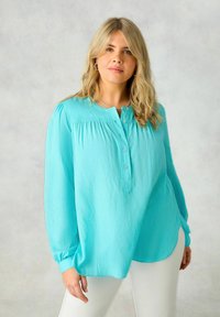 Camicia bottonata azzurro chiaro con maniche lunghe, dettaglio plissettato sul davanti e vestibilità ampia, abbinata a pantaloni bianchi.