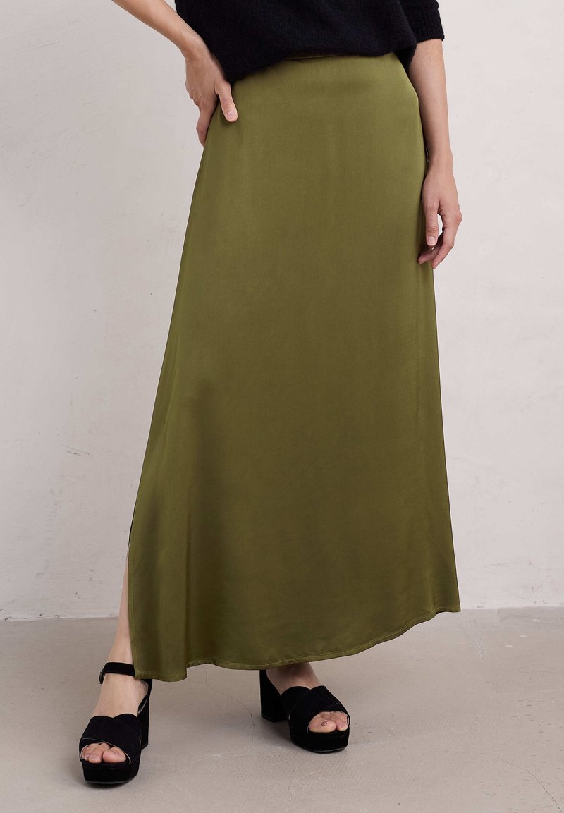 Olivově zelená saténová maxi sukně s rovnou siluetou a bočním rozparkem, v kombinaci s černými semišovými platformovými sandály.