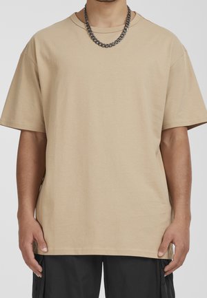 Mies, joka on pukeutunut ylisuureen beigeen t-paitaan, mustiin housuihin ja paksuun hopeaketjuun, seisoo yksinkertaista vaaleataustaa vasten.