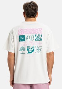 Hvid bomulds t-shirt med et stort flerfarvet grafik på bagsiden, der indeholder designs som træer, hoveder og abstrakte former.