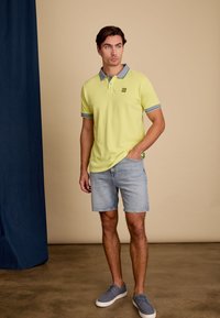 Polo vert lime avec col et poignets gris, associé à un short en denim clair et des chaussures bleues. Détail de logo simple sur la poitrine.
