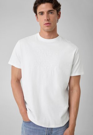 Jeune homme aux cheveux courts et bouclés, portant un t-shirt blanc uni à manches courtes et un jean bleu clair, debout devant un fond gris.