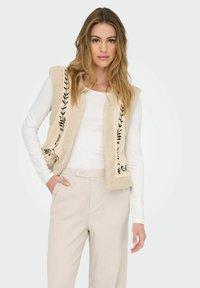 Beige sherpa-väst med svart bladsbroderi längs kanterna, bädd över en vit långärmad topp, kombinerat med ljusbeiga byxor.