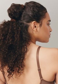 Braune, lockige Haare sind zu einem hohen Dutt mit einem Haargummi gestylt, der die strukturierten Locken zur Geltung bringt. Das Motiv trägt kleine Creolen und ein braunes Trägershirt.