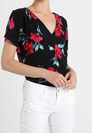 Czarny kwiatowy top z dekoltem w serek, krótkimi rękawami oraz żywym czerwono-turkusowym wzorem kwiatowym. W połączeniu z białymi spodniami.