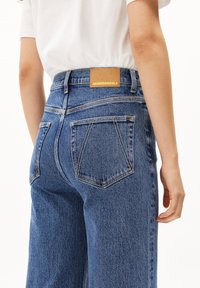 Jeans de mezclilla azul de cintura alta con corte recto, que cuentan con cinco bolsillos, detalles de costura y una etiqueta de cuero marrón en la cinturilla.