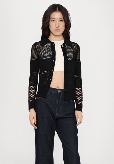 Cardigan nero con design a maglia aperta, dotato di maniche lunghe, colletto e chiusura con bottoni. Indossato sopra un top cropped color crema.