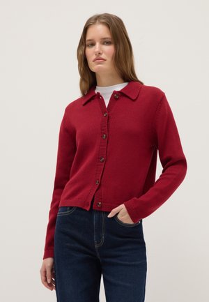 Jonge vrouw draagt een aansluitend rood vest met knopen en kraag, wit ondershirt en donkerblauwe spijkerbroek, staand met één hand in haar zak.