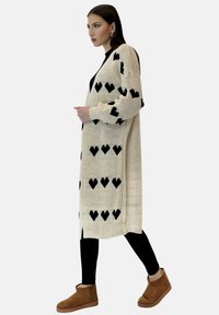 Knitade koftor i kräm med svarta hjärt-mönster, långa ärmar, öppen front och sidoflikar. Matchad med svarta leggings och bruna ankelboots.