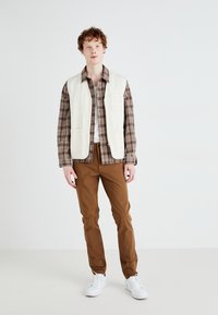 Lett, kremfarget vest over en brun rutete skjorte, paret med brune bukser og hvite sneakers. Glatt stoff og avslappet passform.