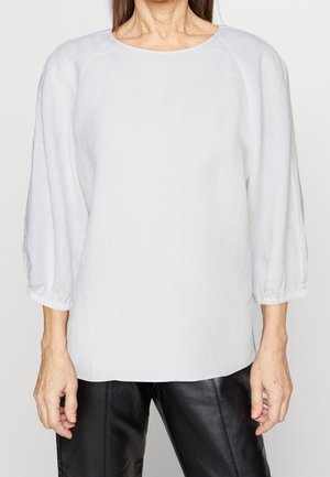 Vrouw die een losse witte blouse met lange mouwen en elastische manchetten draagt en zwarte leren broek, afgebeeld van nek tot heupen tegen een effen achtergrond.