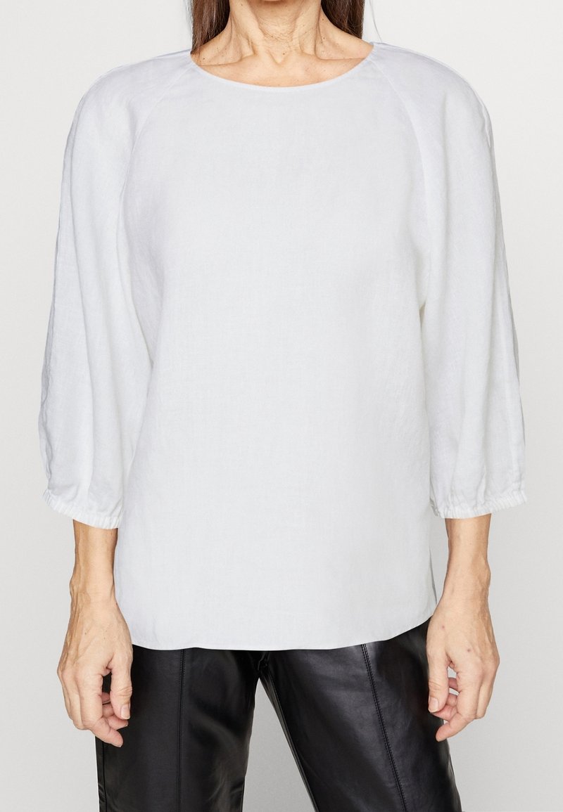 Femme portant un chemisier blanc ample à manches longues avec des poignets élastiques et un pantalon en cuir noir, montrée du cou aux hanches sur un fond uni.