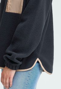 Svart fleece genser med tan nylon brystlapp. Har et avrundet skjær og elestisk mansjett, som fremhever en teksturert overflate.