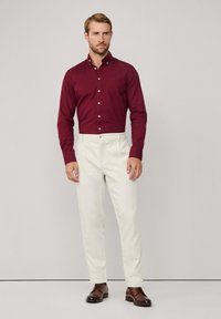 Camicia a maniche lunghe bordeaux con bottoni abbinata a pantaloni color crema e scarpe marrone scuro. La camicia presenta un piccolo logo sul petto.
