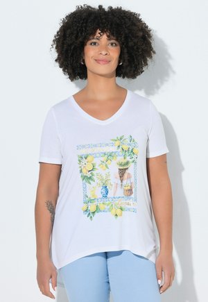 Donna con capelli ricci che indossa una maglietta bianca con scollo a V con stampa di limoni e motivi botanici, abbinata a pantaloni azzurri, in piedi su sfondo bianco.