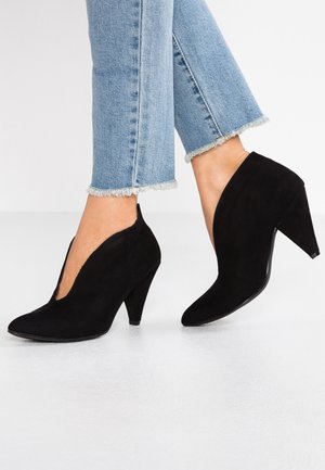 Vrouw draagt zwarte suède enkellaarsjes met puntige neus en middelhoog hakje, gecombineerd met lichtblauwe jeans met afgeknipte zoom, staande op een witte vloer.