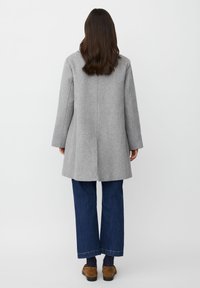 Kvinna med långt brunt hår bär en ljusgrå knälång kappa, mörkblå jeans och bruna skor medan hon står mot en vit bakgrund.