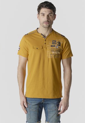 Hombre de pie mirando al frente, vistiendo una camiseta de manga corta de color mostaza con texto impreso y pantalones cortos cargo de mezclilla azul.