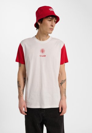 Jonge man die een wit T-shirt met rode mouwen en logo draagt, een rode vissershoed, zwarte broek en zichtbare armtattoeages, kijkt naar rechts.