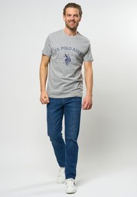 Graues Baumwoll-T-Shirt mit navyfarbenem aufgedrucktem Logo und Text. Normale Passform, kurze Ärmel. Kombiniert mit blauen Jeans und weißen Sportschuhen.