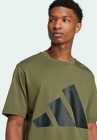 Olivovo zelené bavlnené tričko s veľkým čiernym grafickým logom na prednej strane. Krátke rukávy a okrúhly výstrih. Mäkkučká textura.