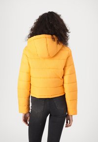 Veste doudoune jaune avec capuche, présentant des sections matelassées horizontales et une coupe courte. Portée sur un jean foncé, avec une texture lisse.