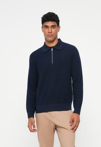 Pullover tricotat de culoare bleumarin, cu guler cu fermoar pe un sfert, textură cu nervuri și un mic logo pe pieptul stâng. Asociat cu pantaloni bej.