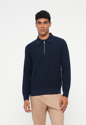 Navyblauwe gebreide pullover met een kwart rits kraag, geribbelde textuur en een klein logo op de linkerkant van de borst. Gecombineerd met beige broeken.