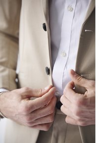 Hender som knepper en beige dressjakke over en hvit skjorte, med fokus på overkroppen og detaljer i klærne.