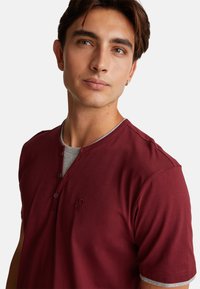 Burgundské tričko s krátkým rukávem henley vyrobené z měkké bavlny, s šedým vnitřním límcem a jemným logem na hrudi.