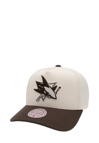 Casquette de couleur crème avec une visière marron. Présente un logo de requin en noir et gris sur le devant. Conçue en tissu avec un design structuré.