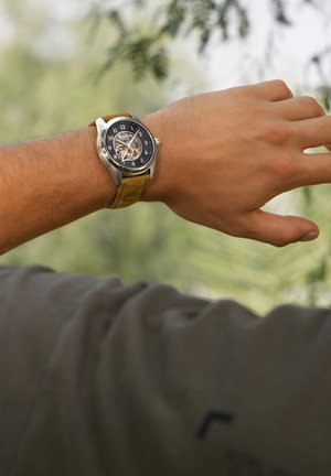 Kairė riešo pusė su rankiniu laikrodžiu, kuriame yra juodas ciferblatas, matomas mechanizmo pavara, sidabrinis korpusas ir rudos odos dirželis su siuvimo detalėmis.