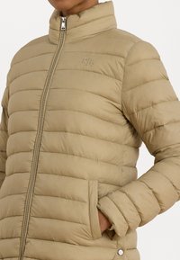 Giacca imbottita color sabbia con colletto alto, caratterizzata da un design a righe verticali, tasche laterali e chiusura frontale con zip, arricchita da un accento con logo.