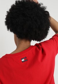 Camiseta de algodón roja con escote en pico y una pequeña etiqueta de marca en la parte trasera con las palabras "TOMMY JEANS" en azul y blanco.