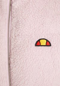 Różowa polarowa kurtka zapinana na zamek z teksturowanej powierzchni, z czarnym, czerwonym i żółtym wyszywanym logo po lewej stronie.