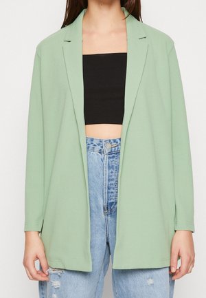 Femme portant un blazer vert clair sur un crop top noir et un jean bleu clair déchiré taille haute, visible des épaules aux hanches.