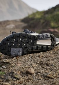 Semelle de chaussure de trail avec un motif en caoutchouc noir et gris reposant sur un sentier montagneux rocheux, avec un arrière-plan flou de verdure.