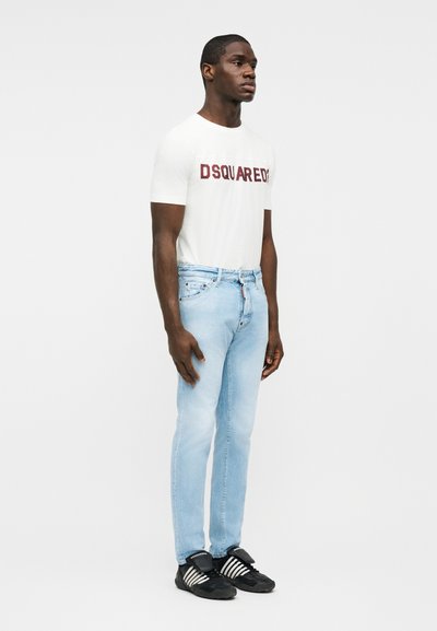 Jeune homme portant un t-shirt blanc avec le logo Dsquared2, un jean bleu clair et des baskets noires à rayures blanches, debout devant un fond blanc.