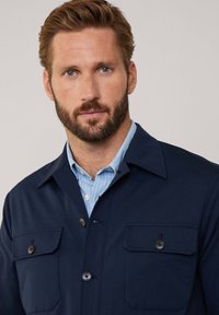 Granatowa koszula z guzikami, z dwiema kieszeniami na piersi, kołnierzykiem i wyraźnie widoczną jasnoniebieską podkoszulką w paski. Gładka tekstura materiału.