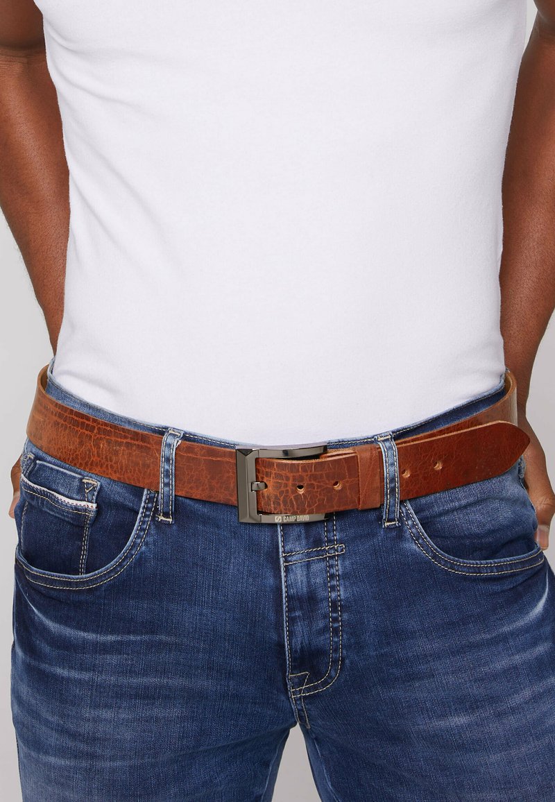 Brown leather belt with a textured finish, featuring a metallic silver buckle, worn with a white t-shirt and blue jeans.