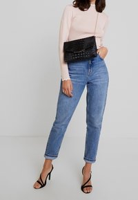 Černé psaníčko se vzorem krokodýlí kůže a zlatým akcentem, pastelově růžový top s dlouhým rukávem a vroubkovanými okraji, a světle modré džíny s vysokým pasem. Černé sandály na podpatku.