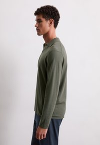 Polo à manches longues de couleur vert olive avec un col classique. Tissu doux, design ajusté. Porté avec un pantalon sombre, présenté de côté.